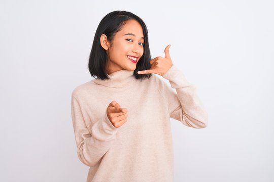 Young chinese woman wearing turtleneck sweater standing over isolated white background smiling doing talking on the telephone gesture and pointing to you. Call me.