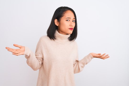Young chinese woman wearing turtleneck sweater standing over isolated white background clueless and confused expression with arms and hands raised. Doubt concept.