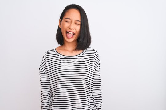Young chinese woman wearing striped t-shirt standing over isolated white background sticking tongue out happy with funny expression. Emotion concept.