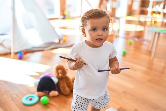 Adorable toddler holding pencils standing around lots of toys at kindergarten