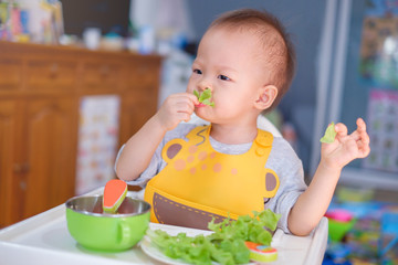 Cute funny little Asian 20 months toddler baby boy child sitting in high chair eating salad,Healthy nutrition and Bio vegetable as solid food for infant. Kid eat vegetables, Eating healthy concept