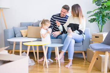 Beautiful family, parents sitting on the sofa drinking coffee looking his kid playing at new home around cardboard boxes