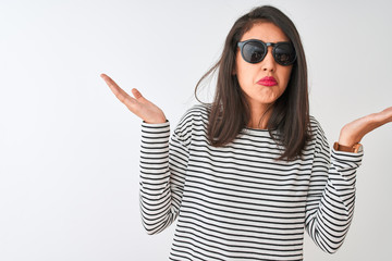 Chinese woman wearing striped t-shirt and sunglasses standing over isolated white background clueless and confused expression with arms and hands raised. Doubt concept.