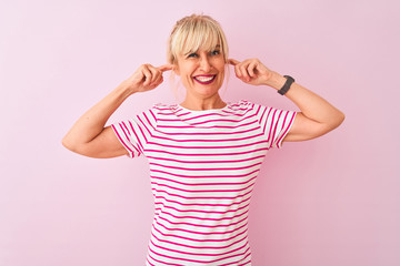 Fototapeta premium Middle age woman wearing striped t-shirt standing over isolated pink background covering ears with fingers with annoyed expression for the noise of loud music. Deaf concept.
