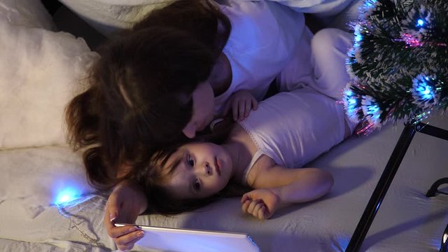 mother and Happy daughter on Christmas evening play on tablet in a children's room in a tent with garlands. baby and mother are playing in room. happy childhood concept.