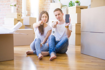 Fototapeta na wymiar Beautiful couple sitting on the floor holding smartphone at new home around cardboard boxes pointing with finger to the camera and to you, hand sign, positive and confident gesture from the front