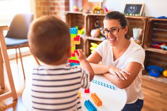 Beautiful teacher and toddler boy playing with construction blocks bulding tower at kindergarten
