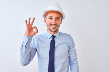 Young business man wearing contractor safety helmet over isolated background smiling positive doing ok sign with hand and fingers. Successful expression.