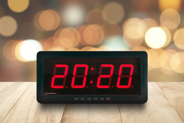 red led light illuminated numbers 2020 on digital electric alarm clock face on brown wooden table top with defocused colorful Christmas lights bokeh background, time symbol for countdown to New Year