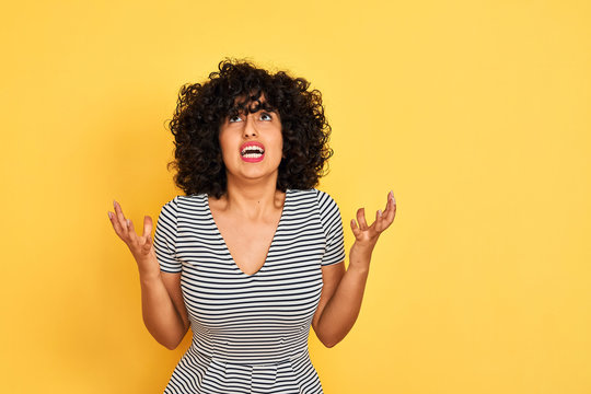 Young arab woman with curly hair wearing striped dress over isolated yellow background crazy and mad shouting and yelling with aggressive expression and arms raised. Frustration concept.