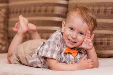 Cute two year old baby in a shirt and a bow-tie is lying on his stomach, leaning on his hands and looking at the camera smiling