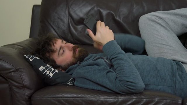 Casual Young Man Lying on Leather Sofa and Using Smartphone