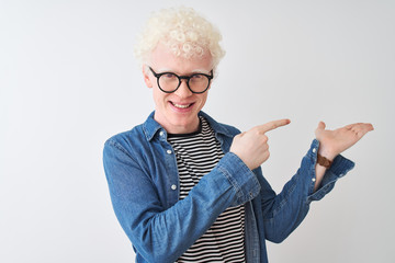 Young albino blond man wearing denim shirt and glasses over isolated white background amazed and smiling to the camera while presenting with hand and pointing with finger.