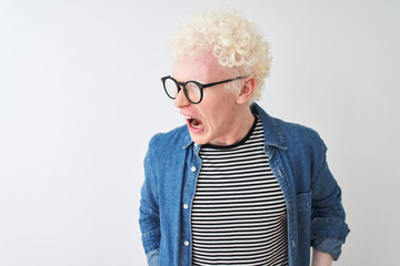 Young albino blond man wearing denim shirt and glasses over isolated white background angry and mad screaming frustrated and furious, shouting with anger. Rage and aggressive concept.