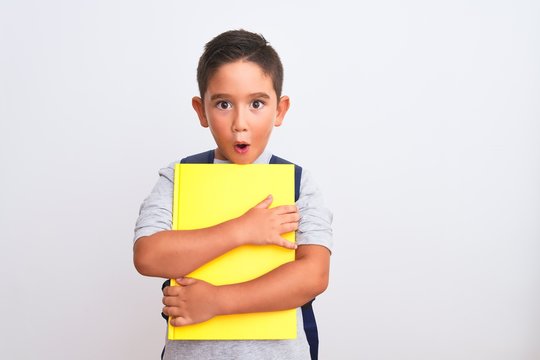 Beautiful student kid boy wearing backpack holding book over isolated white background scared in shock with a surprise face, afraid and excited with fear expression