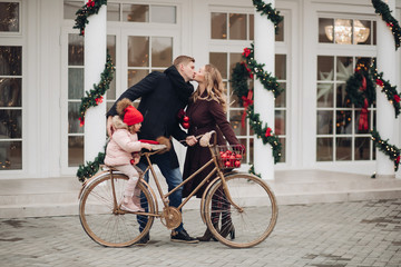 Fototapeta na wymiar Stock photo of loving husband and wife kissing while holding a golden bicycle with their little daughter in red hait on it. They are kissing against beautifully decorated white house with Christmas