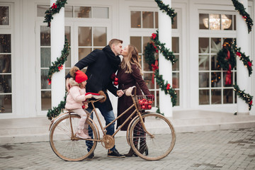 Fototapeta premium Winter family feeling love posing on vintage bicycle at Christmas outdoor decoration background. Happy man and pregnancy woman kissing during cute daughter sitting on bike having positive emotion
