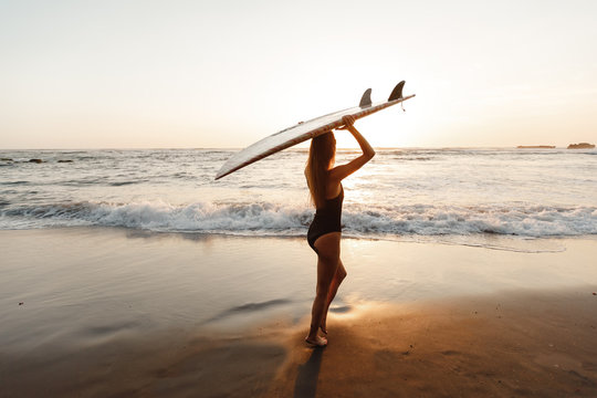 Silhouette of beautiful surfer girl on the beach at sunset. Beautiful young woman with long hair in black  swimsuit with surfboard wait on surf spot at sea beach
