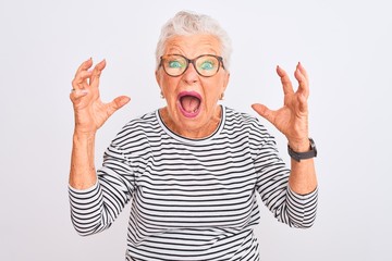 Senior grey-haired woman wearing striped navy t-shirt glasses over isolated white background crazy and mad shouting and yelling with aggressive expression and arms raised. Frustration concept.