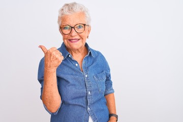 Senior grey-haired woman wearing denim shirt and glasses over isolated white background smiling with happy face looking and pointing to the side with thumb up.