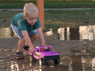 Child (boy, toddler) playing with a pink toy truck in a mud after rain on a flooded playground. Summertime, sunny day, outdoor activity