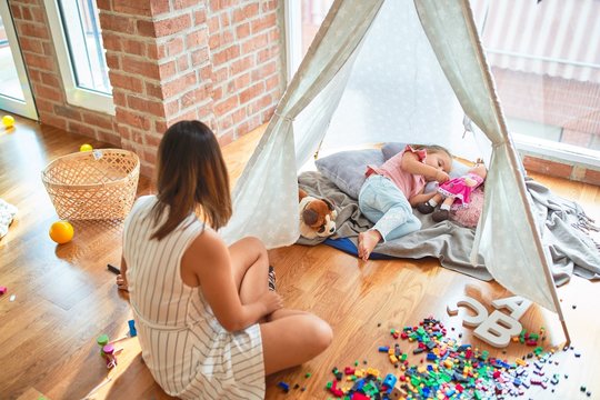 Beautiful teacher and blond toddler girl playing with dolls inside tipi at kindergarten