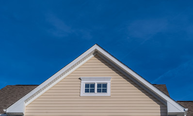 Single gable close up with beige vinyl siding on luxury single family residential home with double attic window in white frame with blue sky background
