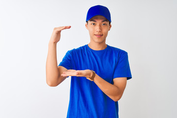 Chinese deliveryman wearing blue t-shirt and cap standing over isolated white background gesturing with hands showing big and large size sign, measure symbol. Smiling looking at the camera. Measuring