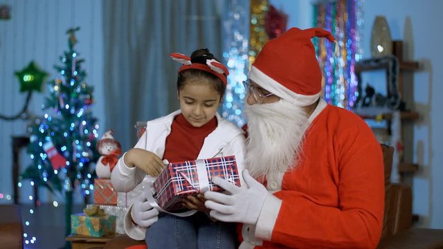 Adorable Indian girl opening her Christmas gift while sitting with happy Santa in India. Young cute kid and old Santa Claus opens a wrapped present together in a decorated room for Christmas celebr...