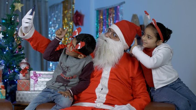 Young cheerful kids happily taking selfies with funny Santa Claus - Christmas time. Happy old Santa clicking pictures with cute little children while enjoying and having fun on Christmas in India