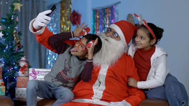 Funny Santa Claus taking selfies with young kids during Christmas celebration in India. Old happy Santa clicking pictures with cute children in different posses - colorful Christmas decorations in ...