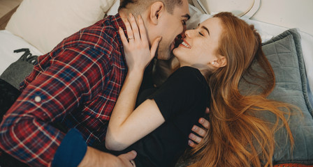 Portrait of a amazing woman with red hair and freckles laughing while her boyfriend is embracing her on the bed trying to kiss.