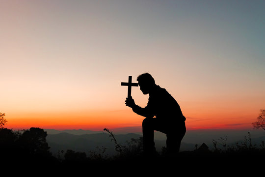 Silhouette human kneeling down praying and holding christian cross for worshipping God at sunset background.Christian, Christianity, Religion copy space background.