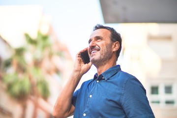 Middle age handsome businessman standing on the street talking on the smartphone smiling