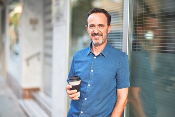 Middle age handsome man leaning on the wall drinking take away coffee smiling