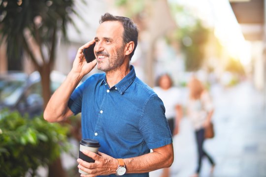 Middle age handsome businessman standing on the street talking on the smartphone drinking take away coffee smiling