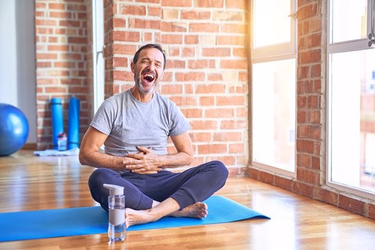 Middle age handsome sportman sitting on mat doing stretching yoga exercise at gym smiling and laughing hard out loud because funny crazy joke with hands on body.