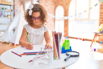 Beautiful toddler wearing glasses sitting drawing using paper and pencils at kindergarten