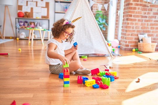Beautiful toddler wearing glasses and unicorn diadem sitting playing with building blocks at kindergarten