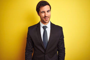Young handsome businessman wearing suit and tie standing over isolated yellow background winking...