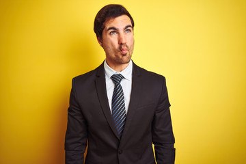 Young handsome businessman wearing suit and tie standing over isolated yellow background making fish face with lips, crazy and comical gesture. Funny expression.