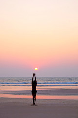 An unrecognizable man standing on a beach arms outstretched to the sun doing a yoga position on an empty beach at sunset