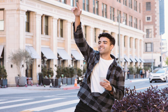 View of a Young Latin Man Hailing a Ride Share Car with a Cell Phone in the City - Afternoon with Bright Lighting