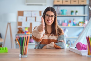 Young beautiful teacher woman wearing sweater and glasses sitting on desk at kindergarten smiling friendly offering handshake as greeting and welcoming. Successful business. - obrazy, fototapety, plakaty