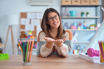 Young beautiful teacher woman wearing sweater and glasses sitting on desk at kindergarten Smiling...