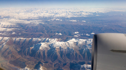 view of sky, snowy mountains, and airplane engine through window while flying