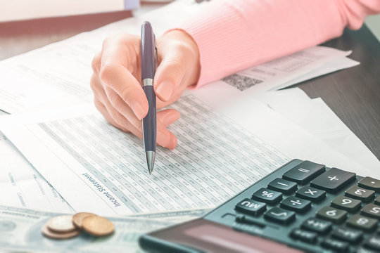 Closeup Side View On The Hand Of Female Accountant Who Is Holding A Pen And Working In An Office With A Stack Of Documents And Tables. Calculation Of Taxes, Profits And Expenses, Saving Money Concept