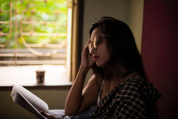 A young and attractive Indian Bengali brunette woman in white sleeping wear with a coffee/tea cup reading book on a bed in front of a window inside in her room. Indian lifestyle.