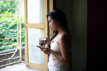 An young and attractive Indian  brunette woman in white sleeping wear standing with a coffee/tea cup and cellphone in front of a window  inside in her room. Indian lifestyle.