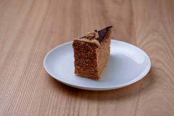 Delicious nut cake glazed with dark chocolate icing served on a white plate on light wooden table background.